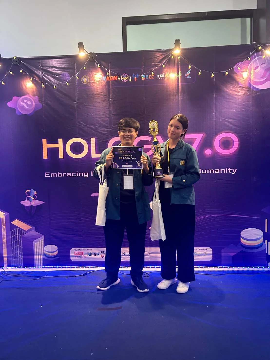 Dua penerima penghargaan membawa plakat dan piala di depan backdrop Hology 7.0