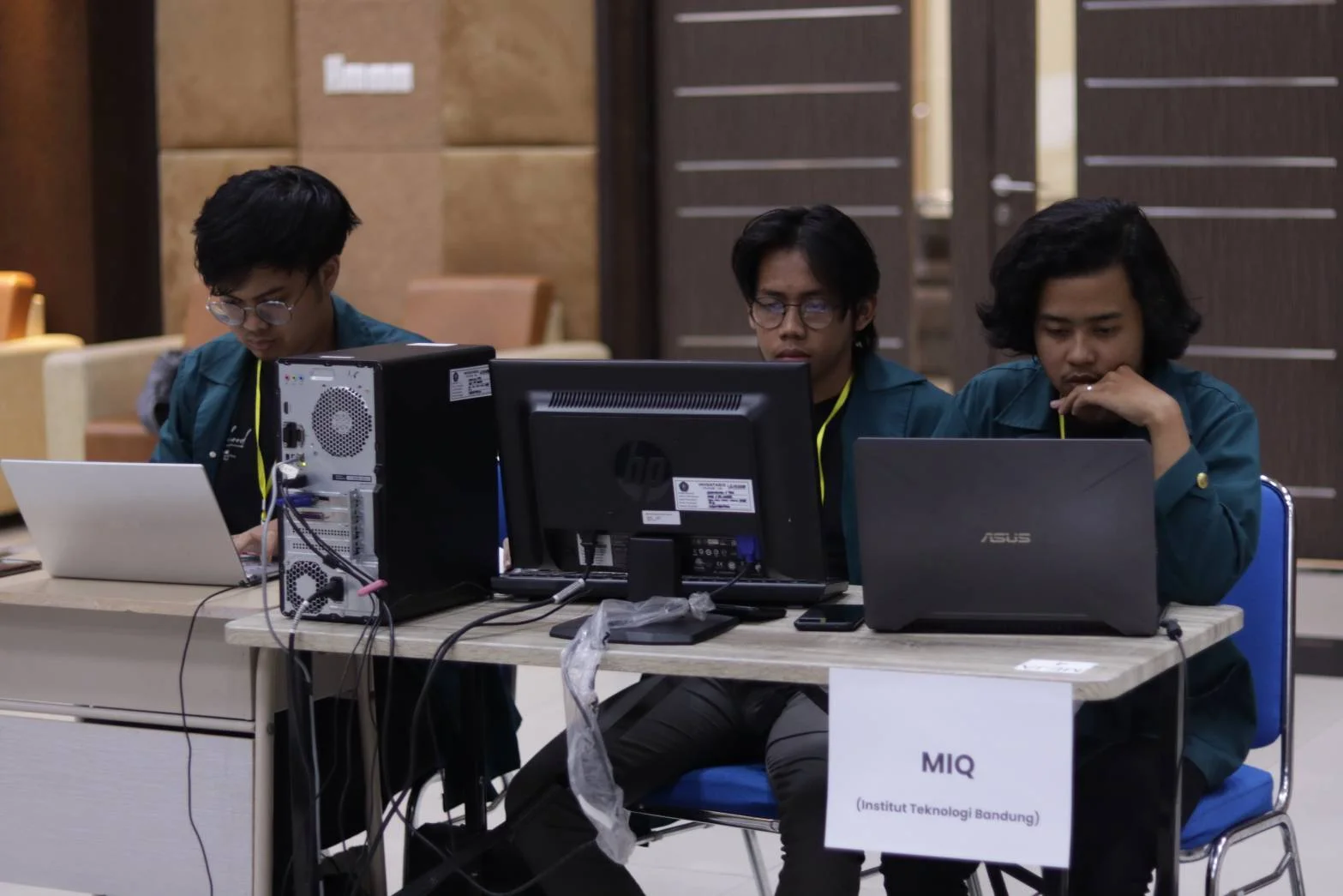 Tiga anggota tim MIQ dari Institut Teknologi Bandung bekerja bersama dengan komputer dan laptop di meja kompetisi